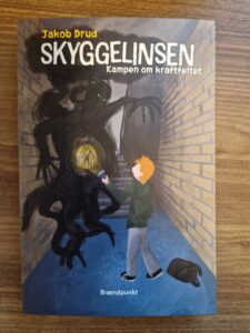 The front page of Skyggelinsen - Kampen om kraftfeltet. It shows a red-haired boy in a green jumper battling a shadow monster in a narrow school hallway.